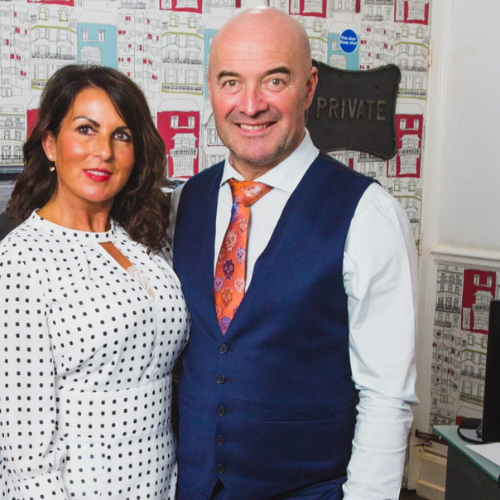 Nina chivers standing in a white dress with black polka dots, standing next Rob Chivers in a blue waistcoat in the Nina Estate Agents office in Barry